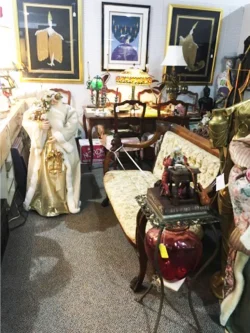 Room filled with antique furniture, framed artwork, lamps, and various decorative vintage items displayed for sale.