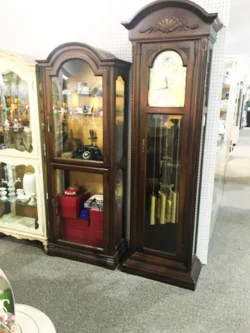 A wooden grandfather clock stands next to a matching display cabinet filled with various items in a store.