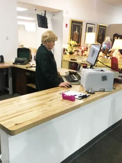 A person stands behind a counter working at a computer in a store with lamps and framed art in the background.