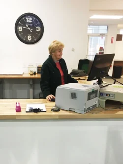 An older woman stands behind a reception desk with a computer, printer, and office supplies; a clock is on the wall.